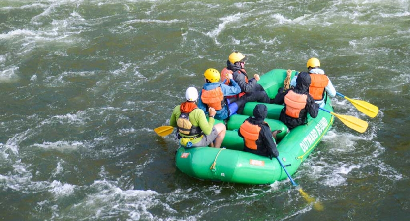 A group of students wearing safety gear paddle a green raft on a river. 